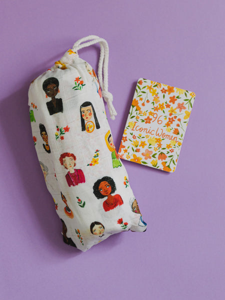 Iconic Women swaddle blanket bundled in matching drawstring bag, sitting alongside a fold-out guide naming all women featured on the print